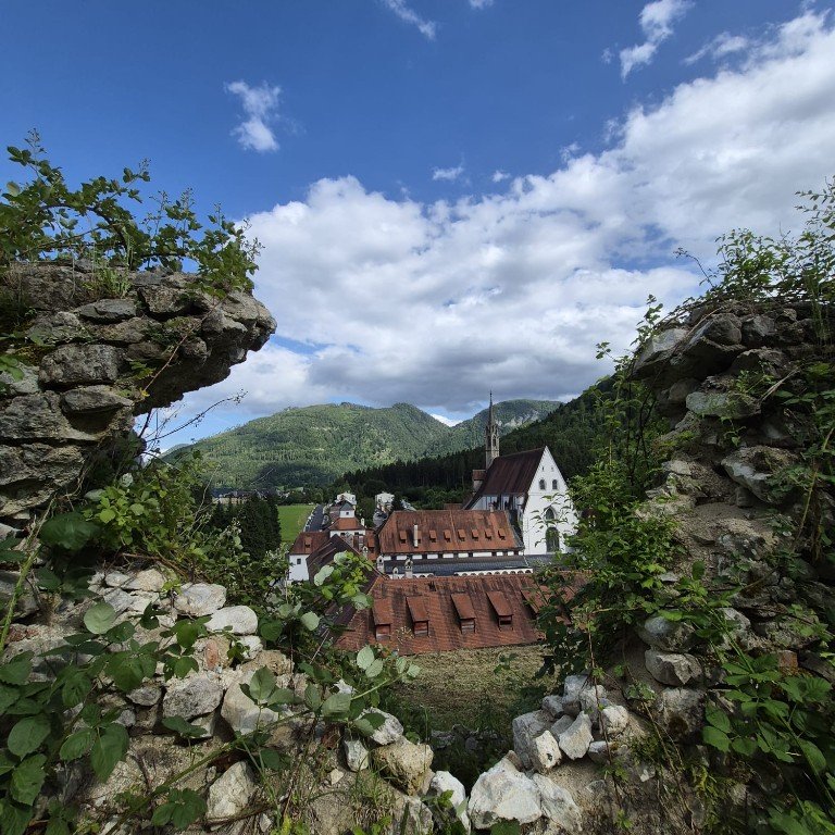 Blick durch eine alte Steinmauer auf eine Dorfkirche in einem bewaldeten Bergtal